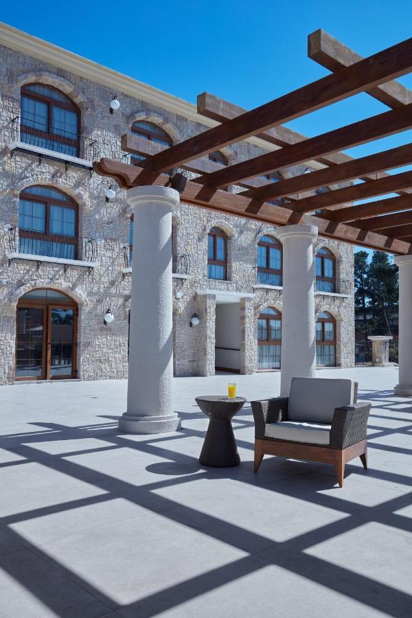 Buona Vitta Gramado Resort & Spa By Gramado Parks Exterior photo The photo shows a well-designed outdoor space with a stone wall building in the background. There are several tall columns that support a wooden pergola overhead, creating a shaded area. In this space, there is a stylish chair and a small table besid
