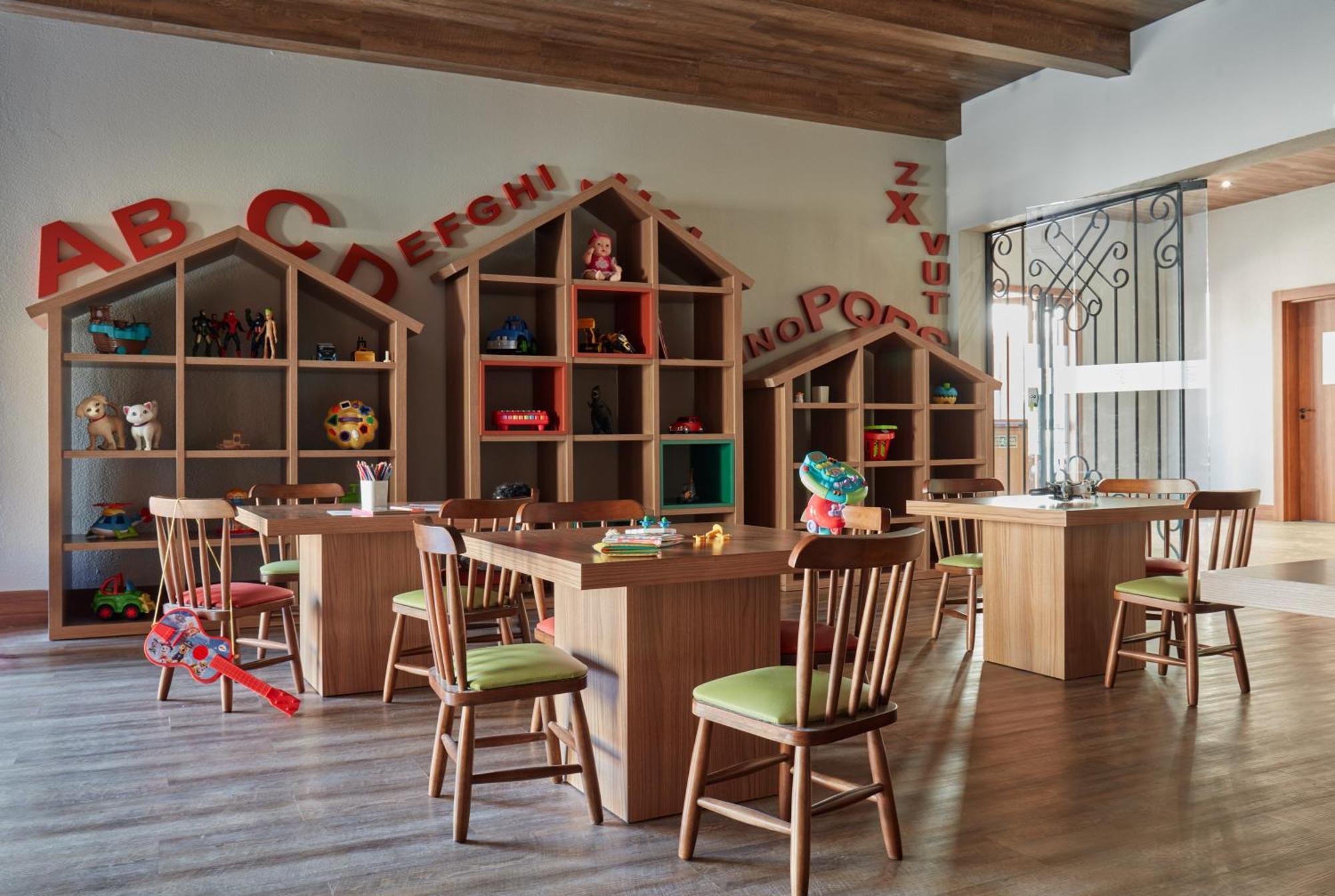 Buona Vitta Gramado Resort & Spa By Gramado Parks Exterior photo The photo shows a classroom or playroom designed for children. It features wooden furniture, including several tables and chairs with green cushions. In the background, there are large, open shelving units shaped like houses, displaying various toys 