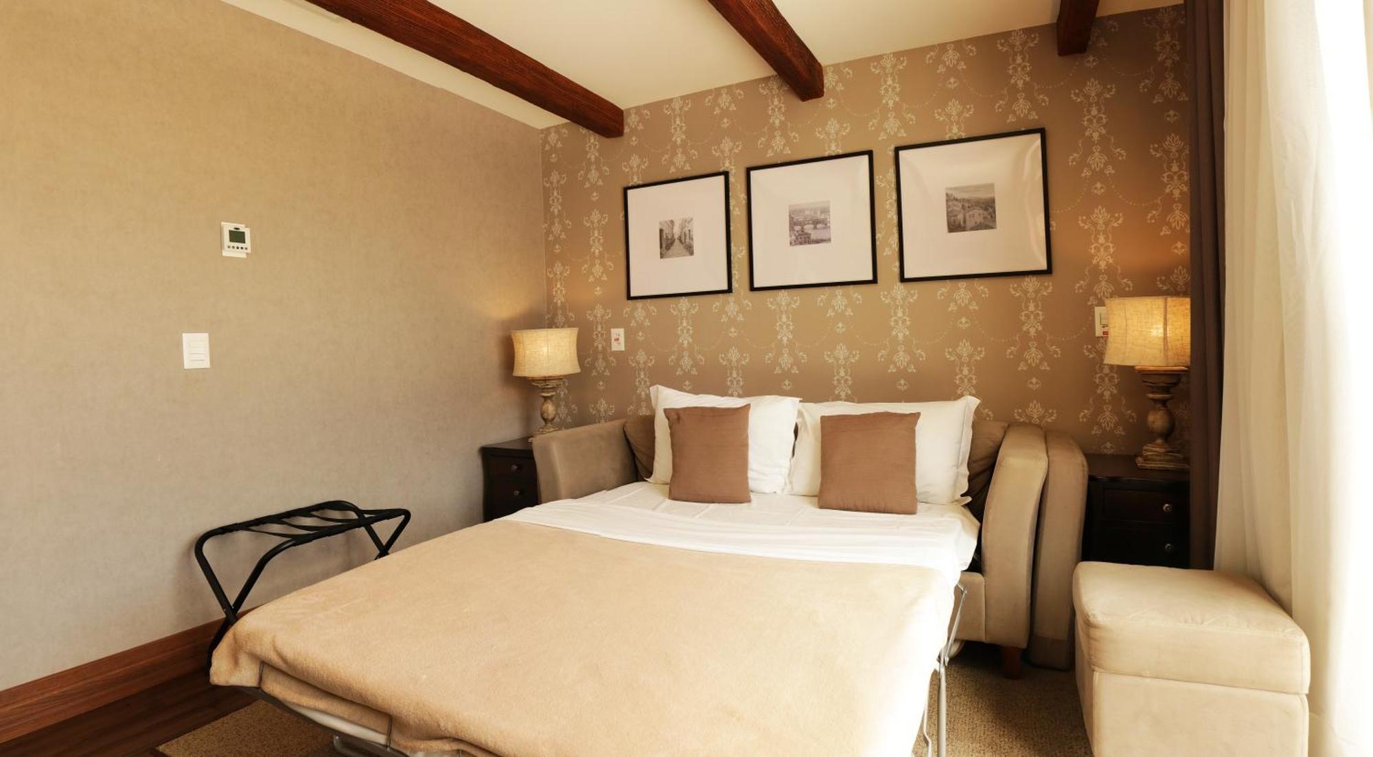 Buona Vitta Gramado Resort & Spa By Gramado Parks Exterior photo The photo shows a cozy bedroom with a neutral color palette. There is a bed with white linens and two brown decorative pillows. On either side of the bed, there are bedside tables with lamps. The walls feature a subtle floral pattern, and three frame