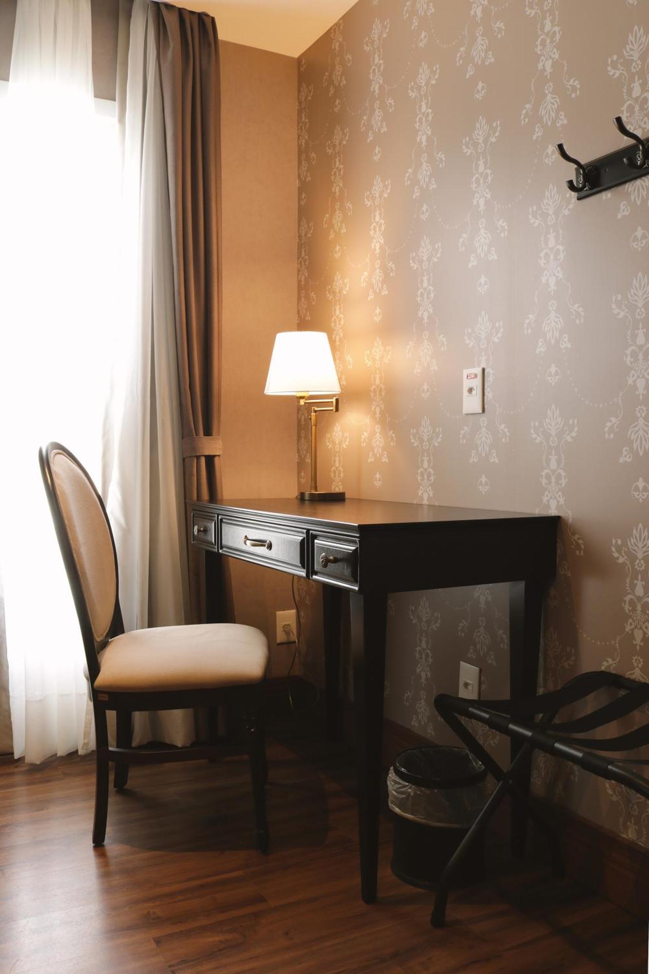Buona Vitta Gramado Resort & Spa By Gramado Parks Exterior photo The photo shows a small desk set against a wall with a decorative pattern. The desk is black with a few drawers and has a small lamp on it. Next to the desk, there is a chair with a light-colored cushion. Soft light is coming through a window, castin
