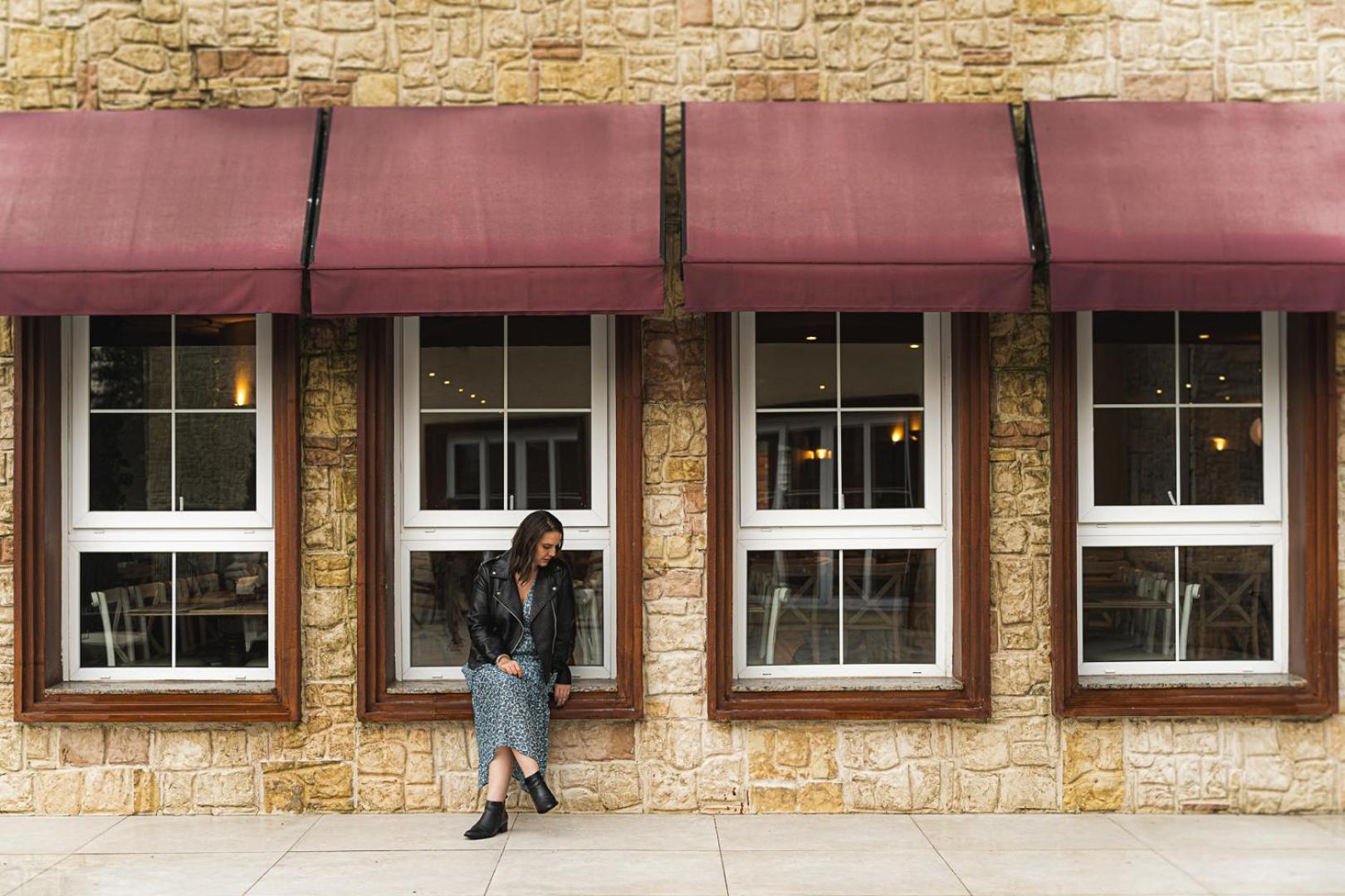 Buona Vitta Gramado Resort & Spa By Gramado Parks Exterior photo The photo shows a woman standing against a stone wall with three large windows in front of her, each framed with wooden borders. The windows have maroon awnings above them. The woman is dressed in a long skirt and leather jacket, leaning slightly aga